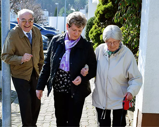 Betreuungskraft begleitet zwei ältere Personen bei einem Spaziergang