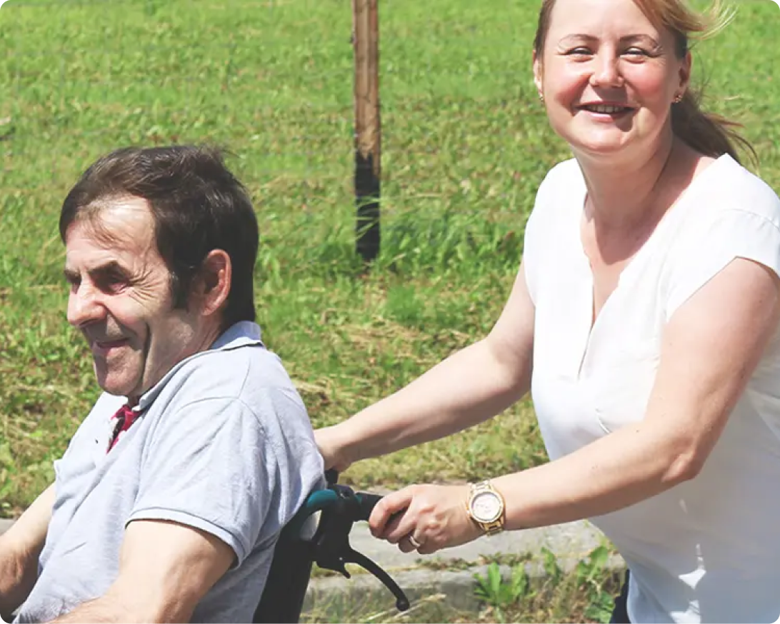 Betreuungskraft begleitet einen Mann im Rollstuhl bei einem Spaziergang im Freien