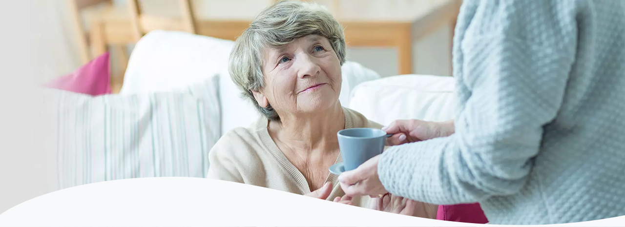 Betreuungskraft reicht einer älteren Frau eine Tasse Getränk zu Hause